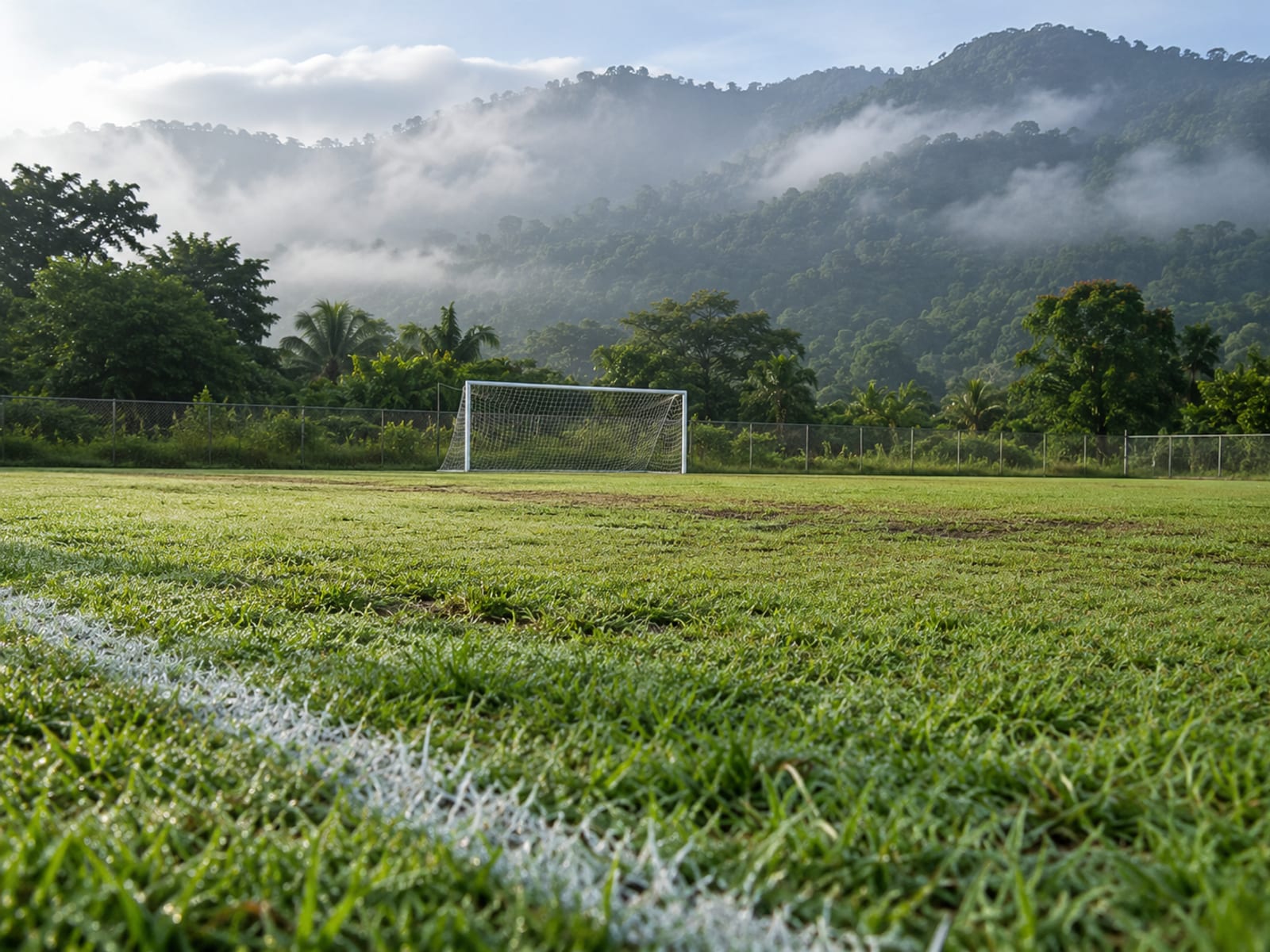 Empty grass pitch with white goalposts at one end and misty green hills beyond