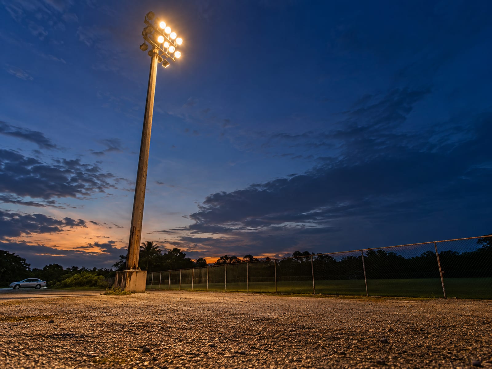Single tall floodlight pole lit against a dusk sky over a gravel car park and dark pitch