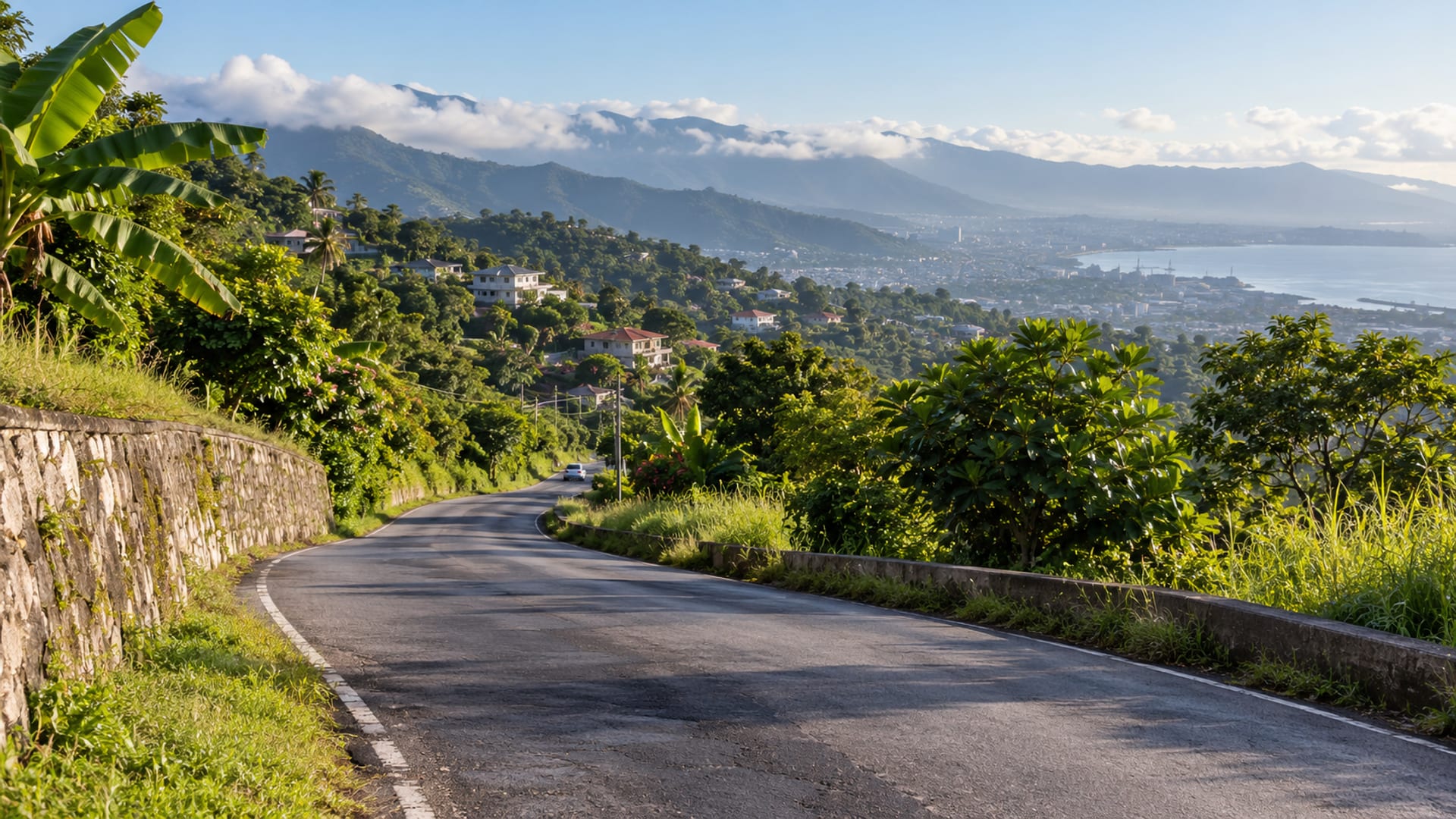 Wide view of a narrow paved road curving down a green hillside in St. Andrew under a pale morning sky, with low cloud over the ridges and a single small plain car far down the road.