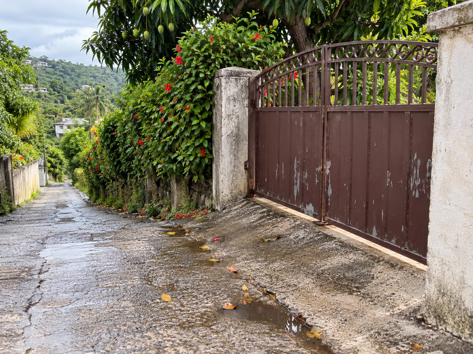 Close view of a closed metal gate and concrete gatepost at the top of a steep narrow lane, with the lane dropping away behind and wet patches on the concrete.
