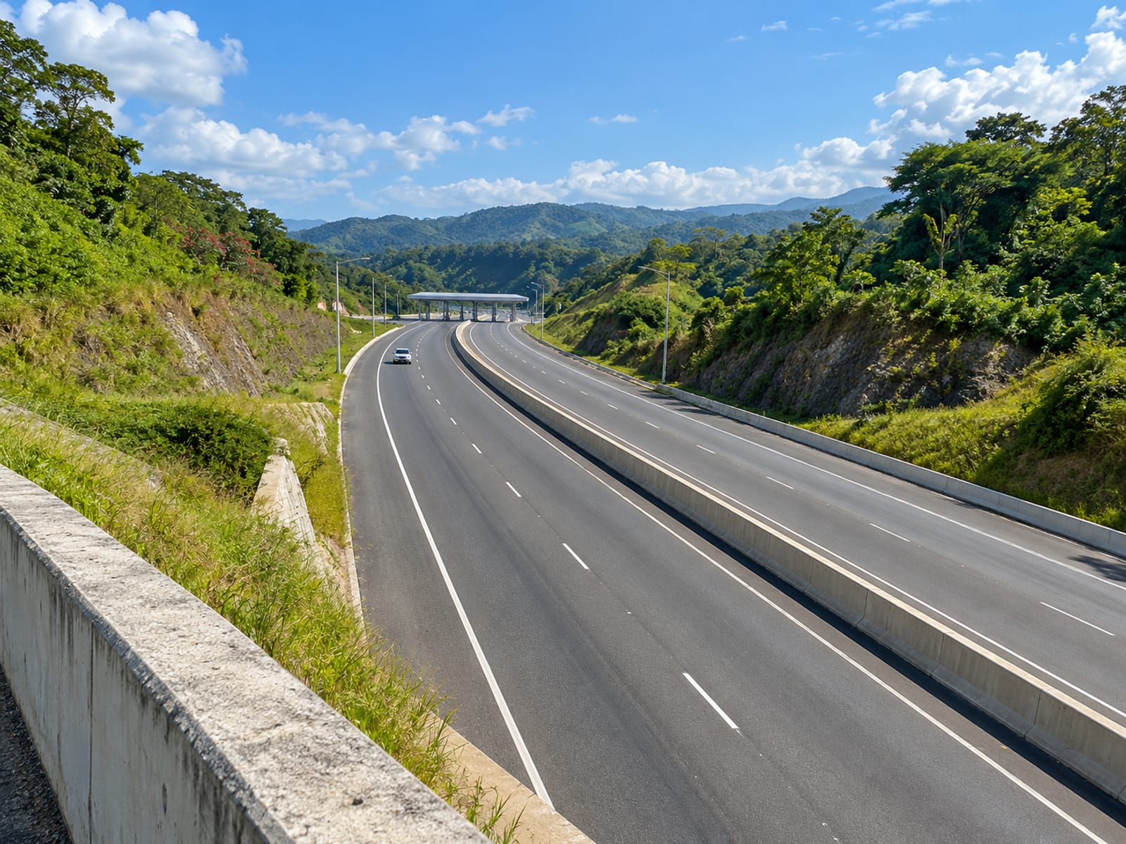 Wide view of a divided highway cutting through green hills, with a plain toll canopy far ahead and a tiny car on the road.