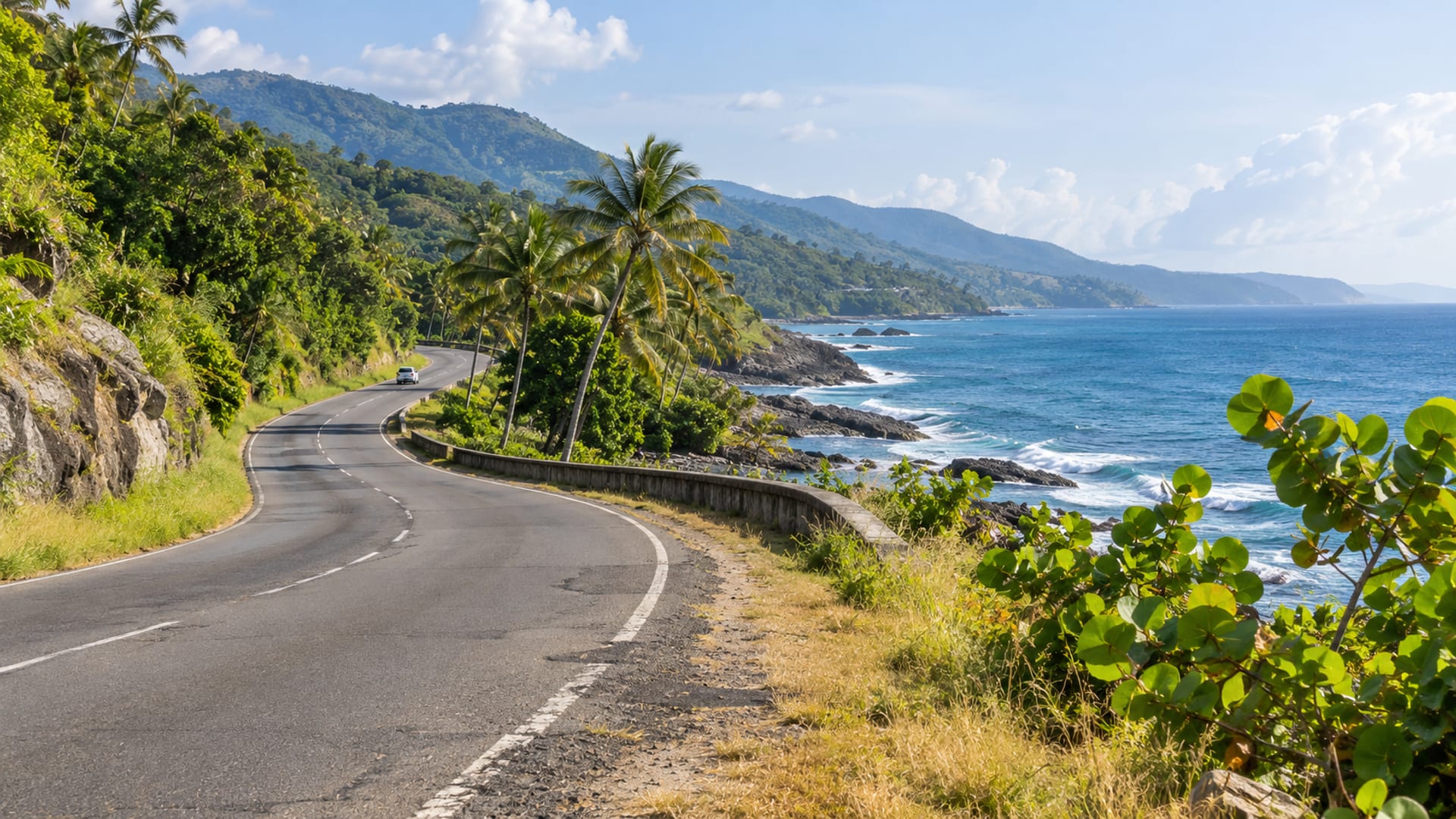 Wide empty coastal road curving beside a calm blue sea with green hills rising inland under a clear sky