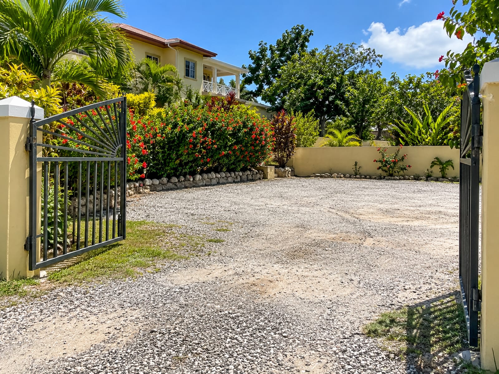 Empty gravel parking area beside a small guesthouse garden with a gate and hibiscus hedge