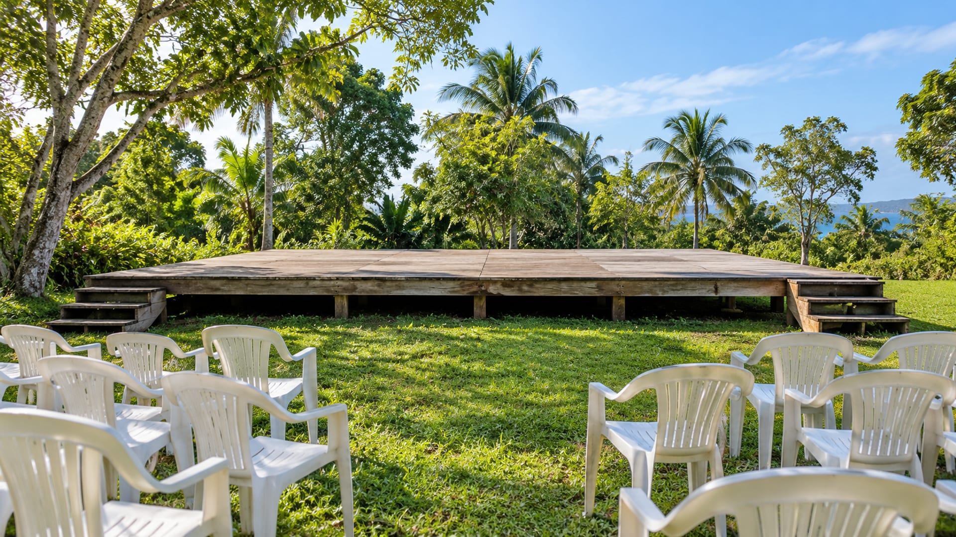 Empty raised timber stage on a green lawn with white chairs and trees behind under a clear sky.