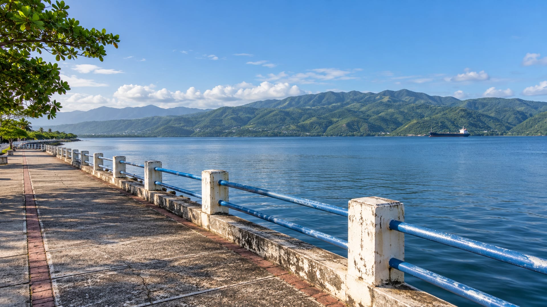 Empty paved harbour promenade with calm water and green hills rising across the bay