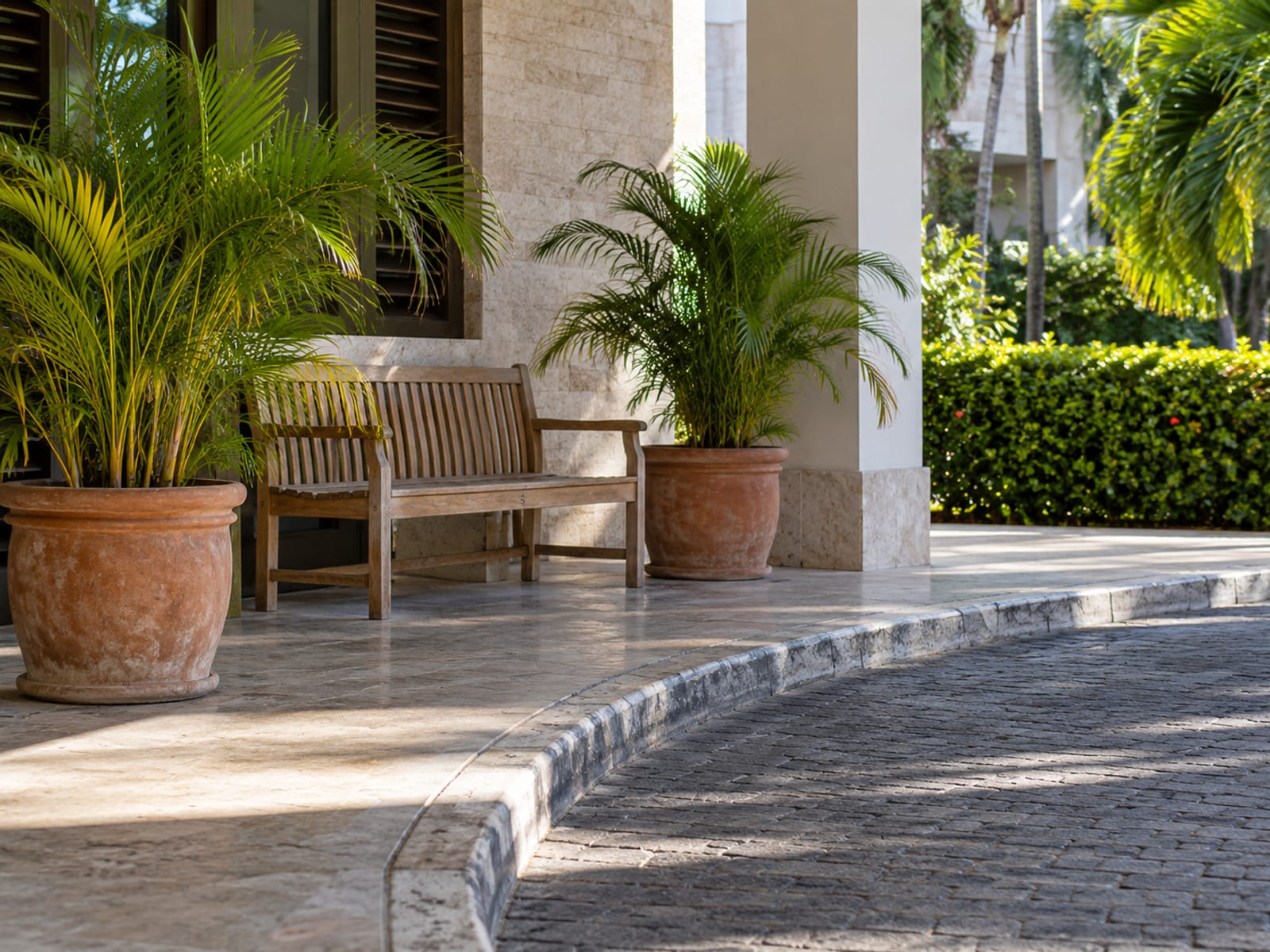 Empty bench and potted palms at a plain hotel entrance kerb with a marked paving edge, no signs