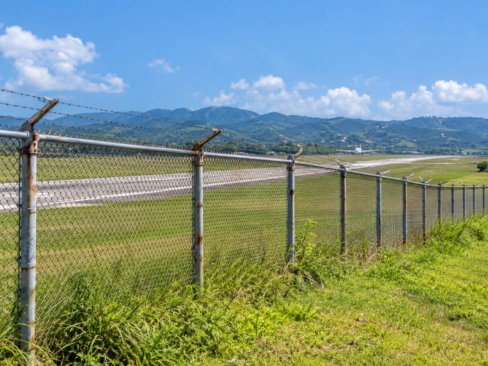 Chain link fence along a grass airfield edge with a small aircraft far away on a runway, no terminal signs