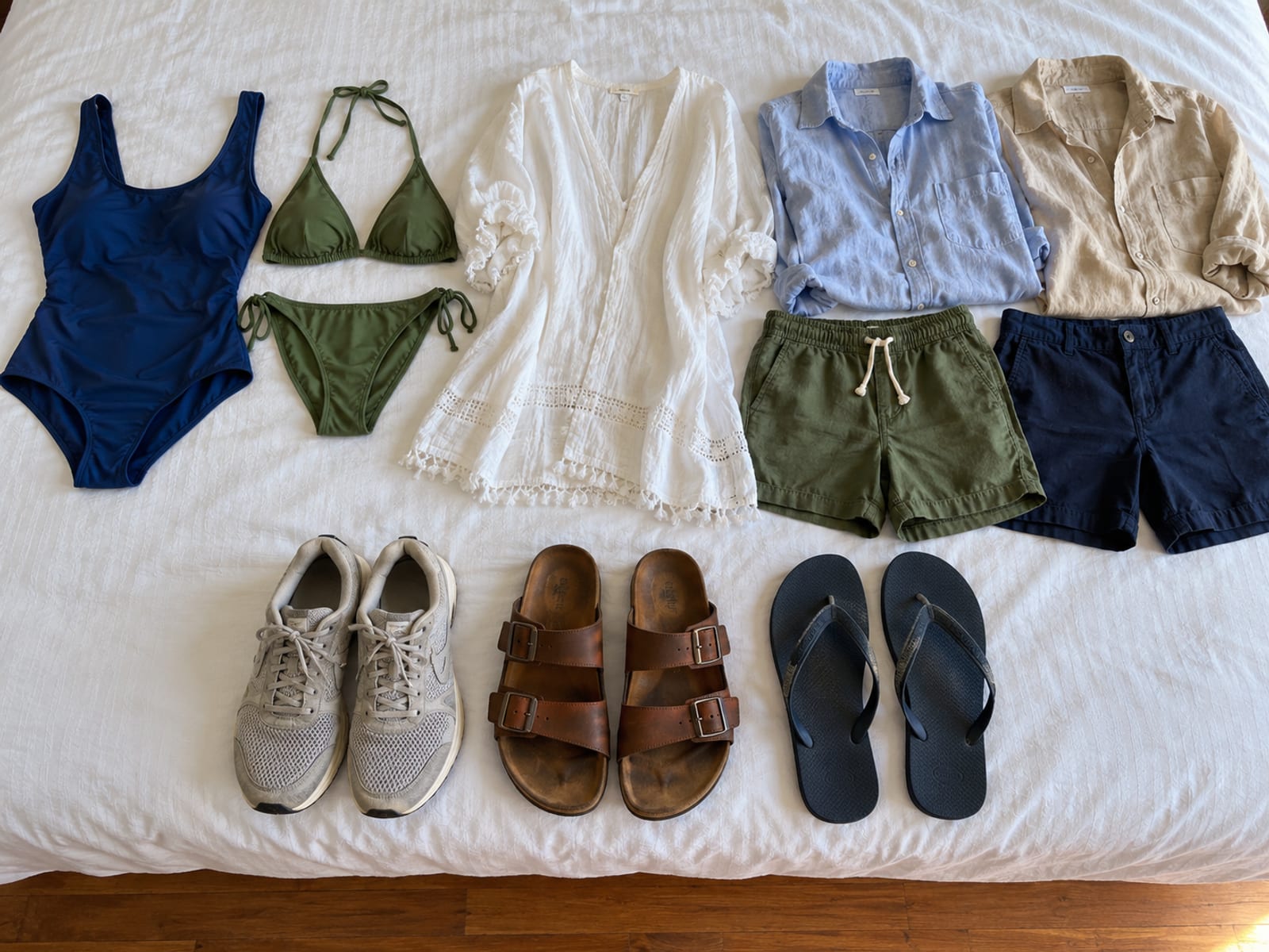 Flat lay on a bed of light cotton and linen clothes, two swimsuits and three pairs of shoes in a row, none with visible brands