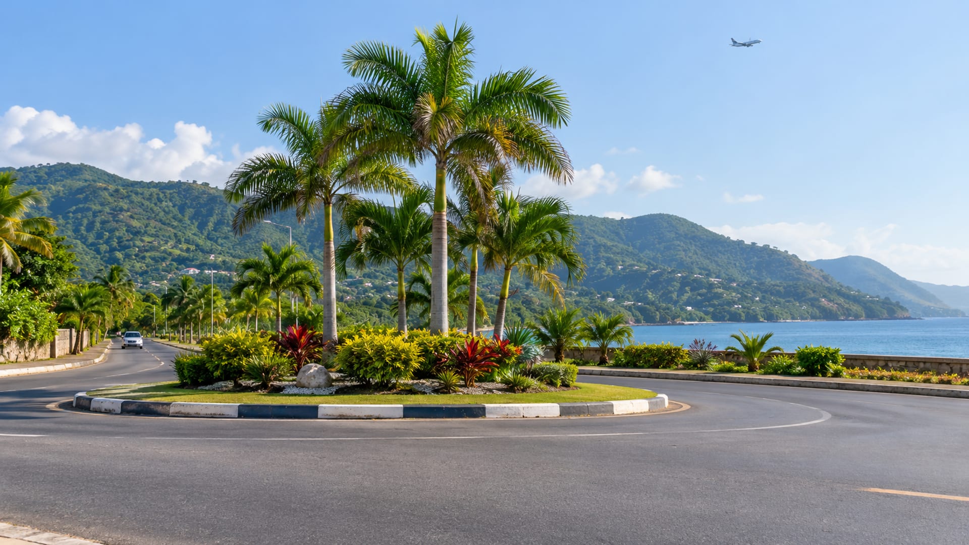 Wide view of a palm planted roundabout island on a coastal road in Jamaica with green hills and a faint plane in the sky