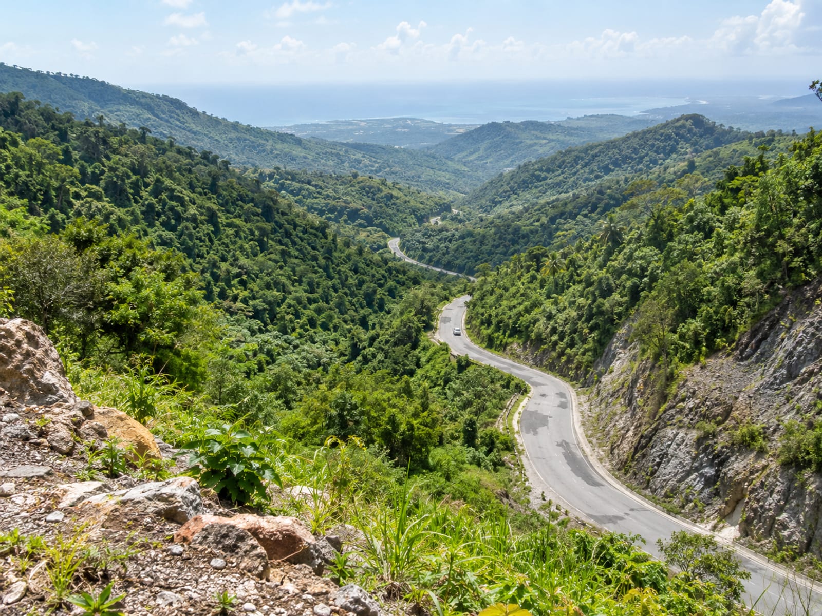 High lookout view of a winding road through green valleys toward a distant sea, with one tiny plain vehicle